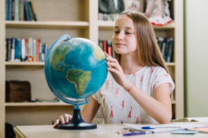 A girl in a light-colored shirt interacts with a globe, surrounded by books on shelves in a study environment.