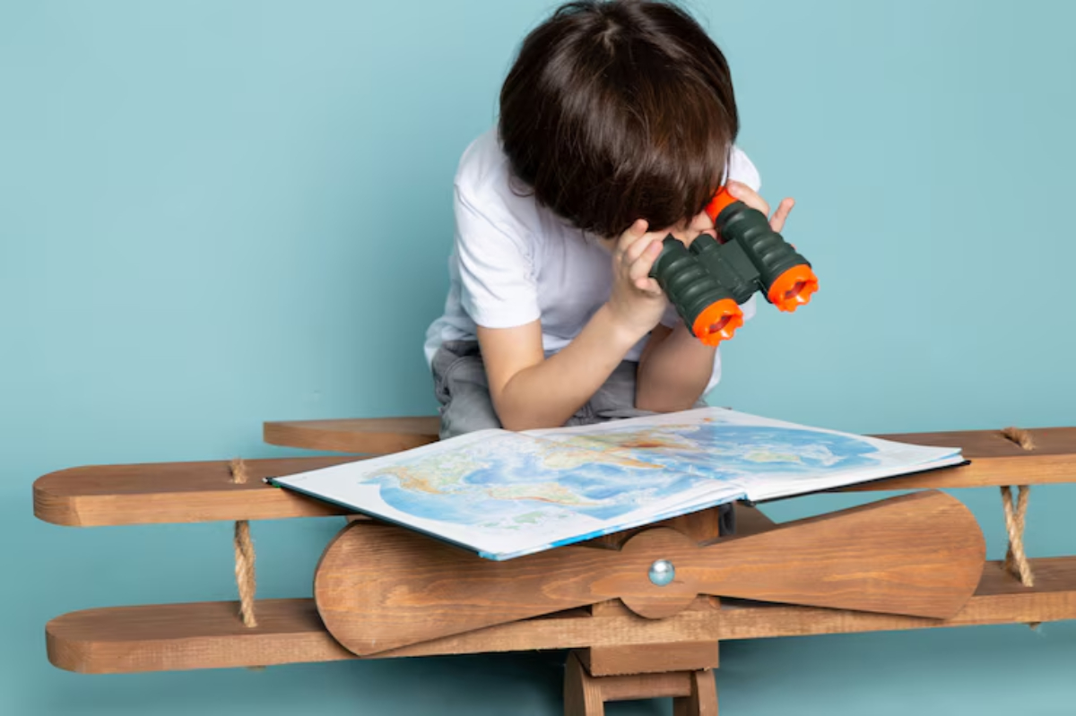 A young boy peers through binoculars while sitting on a wooden airplane toy, examining a colorful map laid out in front of him.