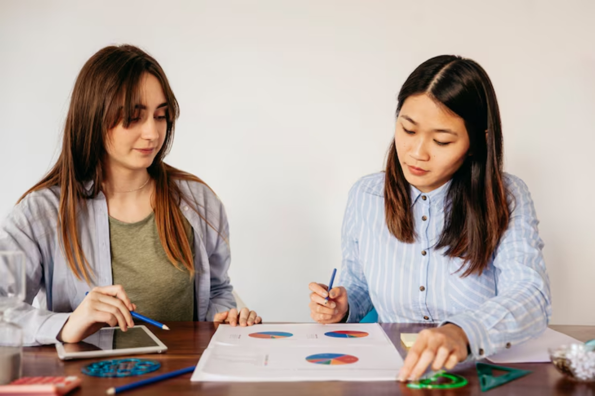 Two women sit at a table, analyzing colorful pie charts and graphs, with stationery and a smartphone nearby.