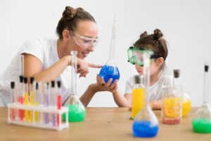 A woman and a girl in goggles explore blue liquid in a flask, surrounded by colorful test tubes and beakers on a table.