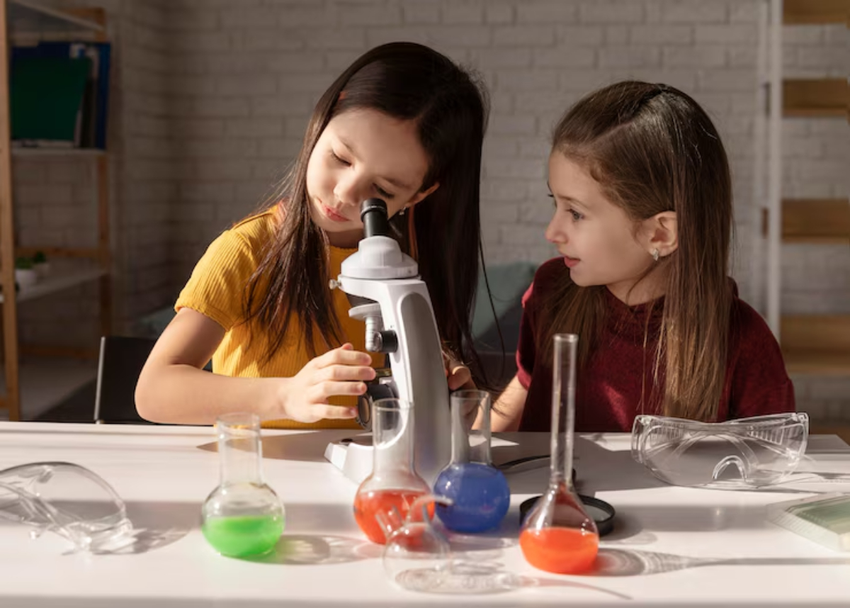 Two children examine samples through a microscope, surrounded by colorful liquids in various laboratory glassware. Safety goggles are nearby.