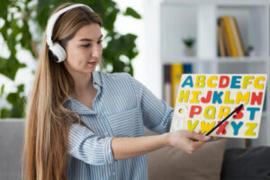 A person wearing headphones holds a colorful alphabet board, pointing at letters with a stick, in a bright, plant-filled room.
