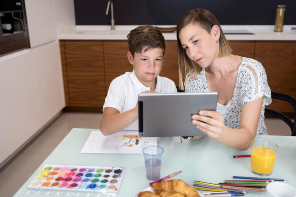 A child and an adult sit at a table, using a tablet together, surrounded by art supplies, juice, and pastries.