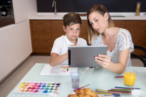 A child and an adult sit at a table, using a tablet together, surrounded by art supplies, juice, and pastries.