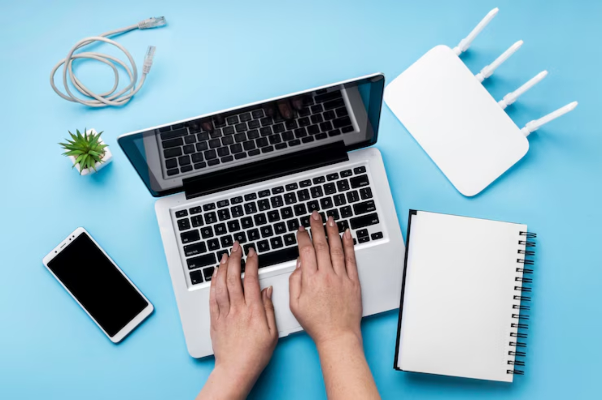 Top view of a laptop, smartphone, notepad, and a Wi-Fi router on a blue surface, with hands typing on the laptop keyboard.