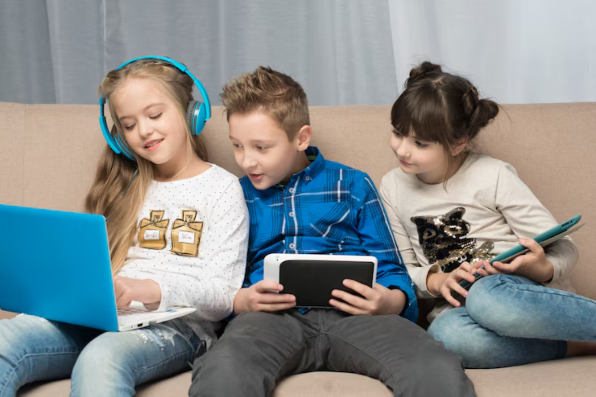 Three children are seated on a couch, engaged with various electronic devices: a laptop, tablet, and phone, in a cozy indoor setting.