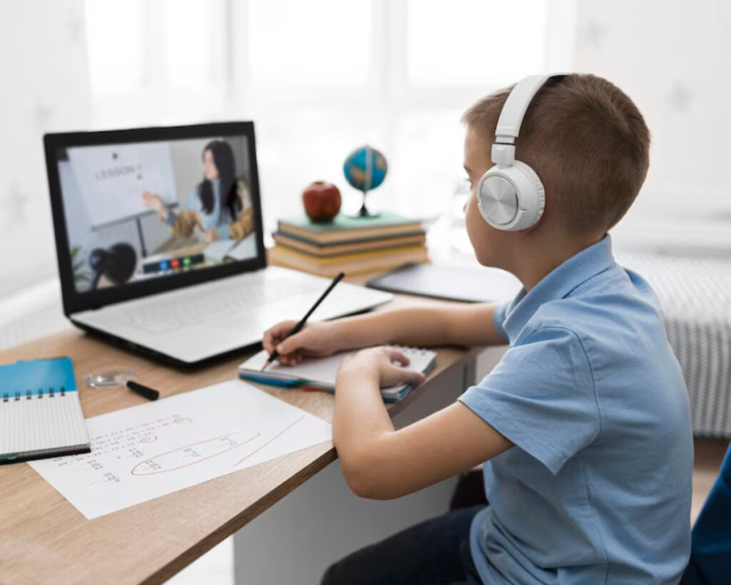 A young boy in headphones attentively participates in an online class, taking notes while seated at a desk with books and papers.
