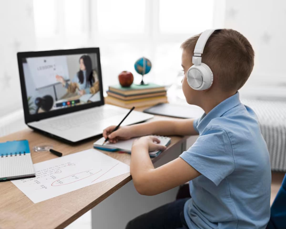 A young boy in headphones attentively participates in an online class, taking notes while seated at a desk with books and papers.