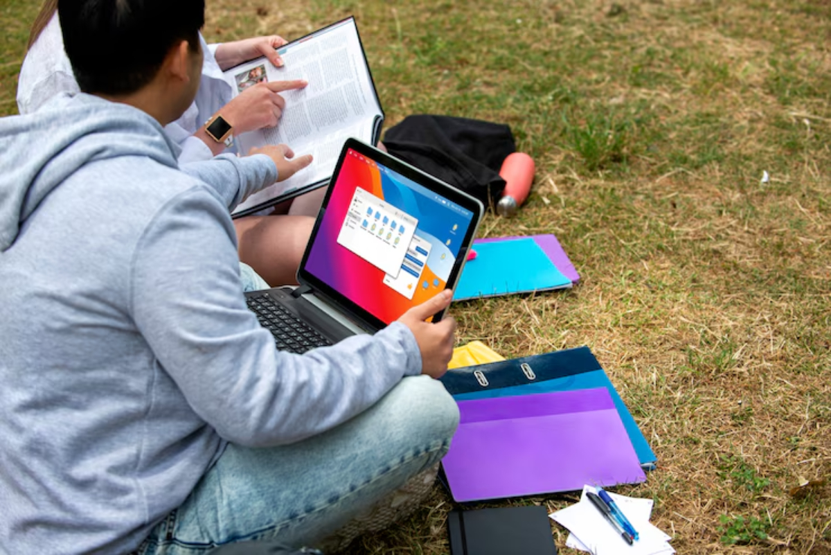 Two students study outdoors, one pointing at a textbook and the other holding a laptop displaying a desktop interface, surrounded by stationery.
