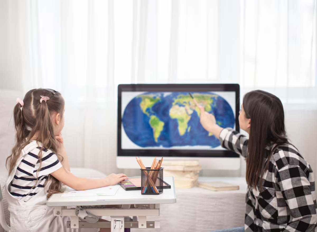 A girl watches as a woman points to a world map on a computer, engaged in learning at a desk with art supplies.