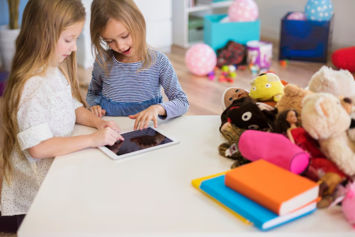 Two young girls engage with a tablet on a table, surrounded by colorful toys and books in a bright playroom setting.