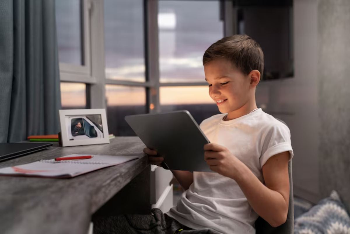 A boy seated at a desk holds a tablet, surrounded by books and a framed photo, with a sunset visible through the window behind him.