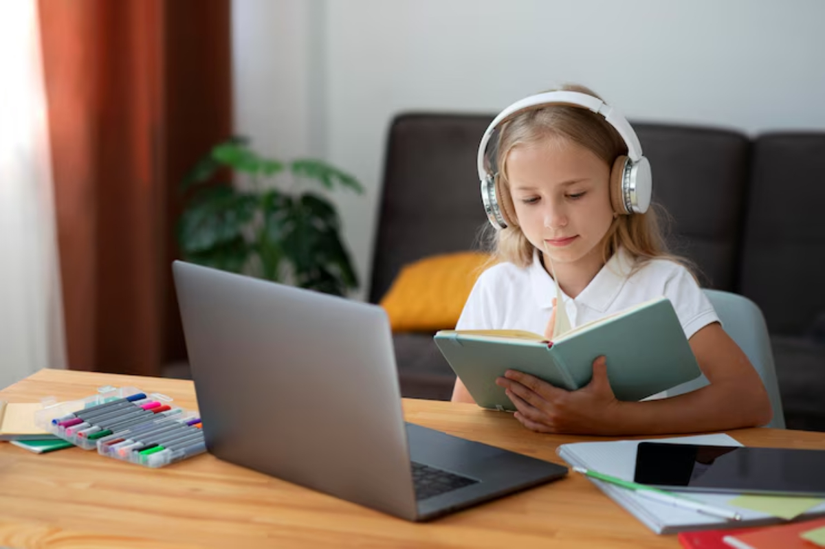 A child wearing headphones sits at a wooden desk, reading a book, with a laptop and colorful markers nearby. A plant is visible in the background.