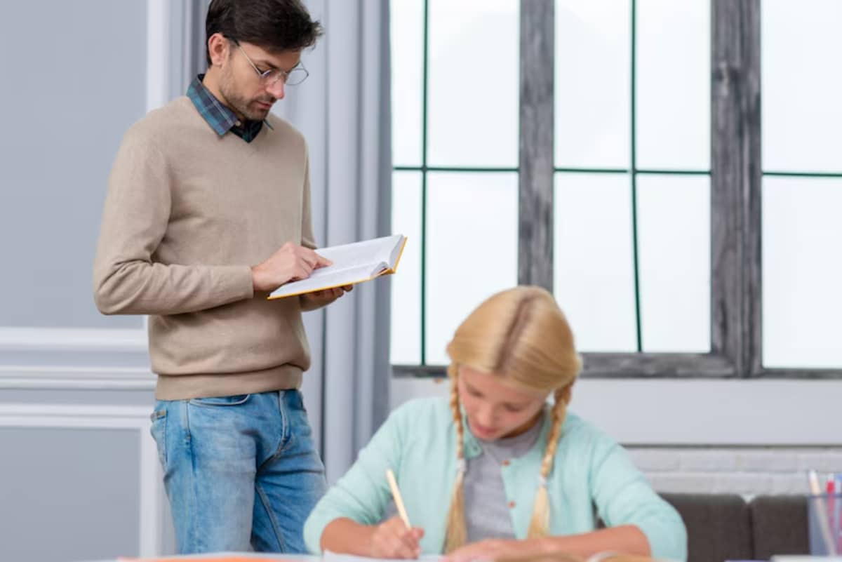 A student sits at a desk writing, while an instructor stands nearby, holding an open book in a well-lit room by large windows.