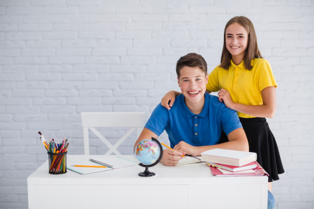 A boy in a blue shirt sits at a desk with a globe, books, and stationery. A girl in a yellow shirt stands behind him, smiling.