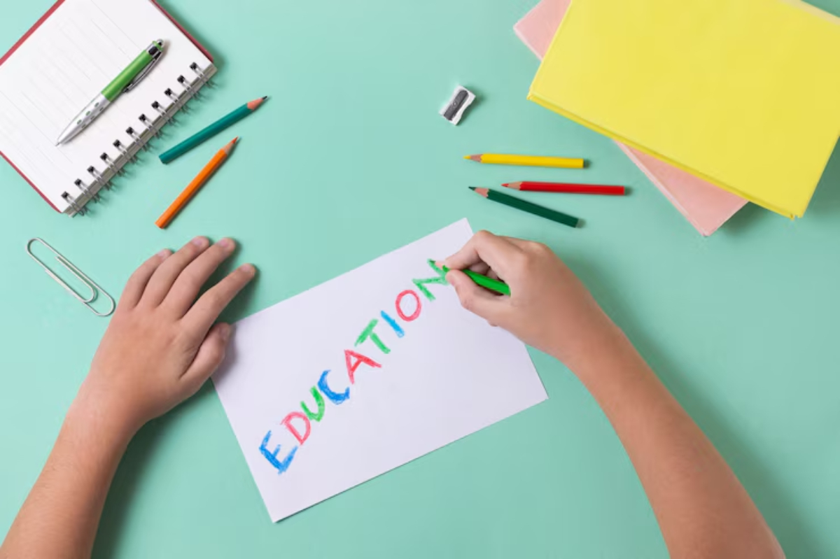 A child's hand writes EDUCATION in colorful letters on a white sheet, surrounded by stationery on a mint green desk.