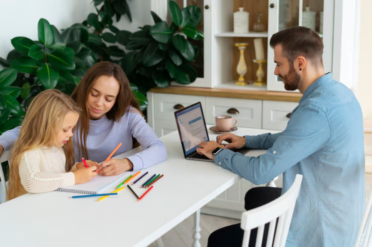 A man works on a laptop at a bright table while two children draw with colored pencils, surrounded by lush indoor plants.