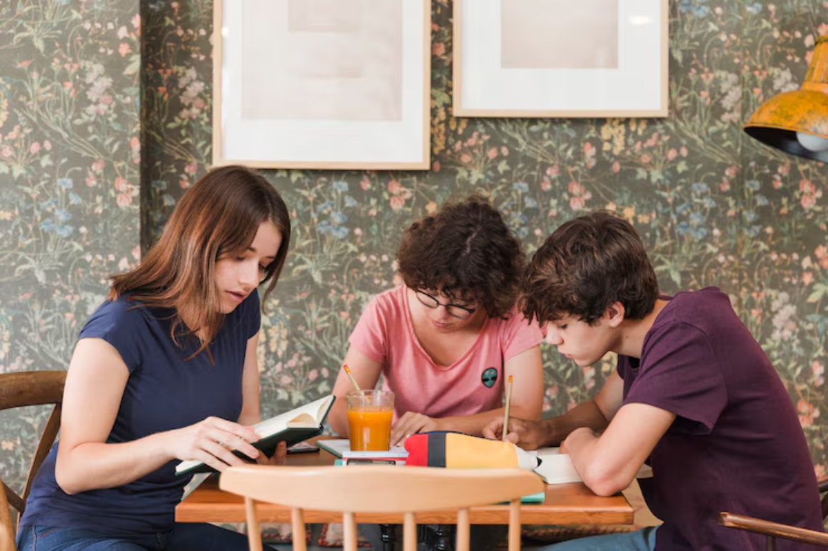 Three individuals seated around a wooden table, engaged in study with books and an orange drink, set against a floral wallpaper backdrop.