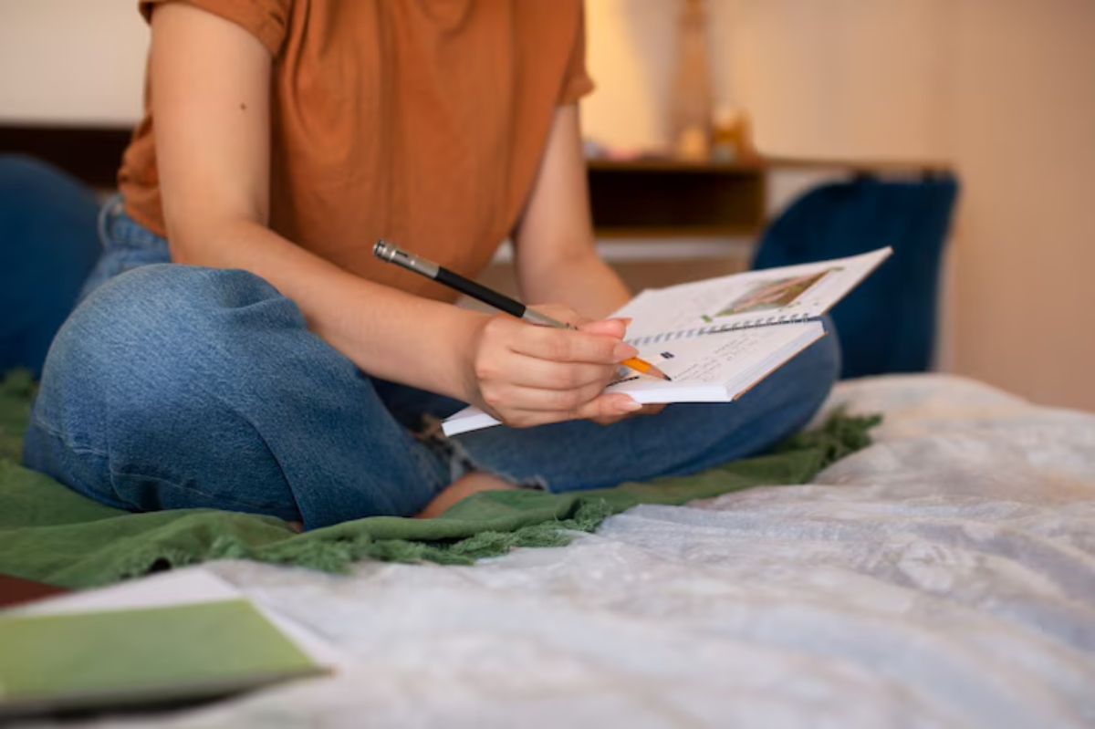 A person sits cross-legged on a bed, writing in a notebook with a pen and orange pencil, surrounded by a cozy, relaxed atmosphere.