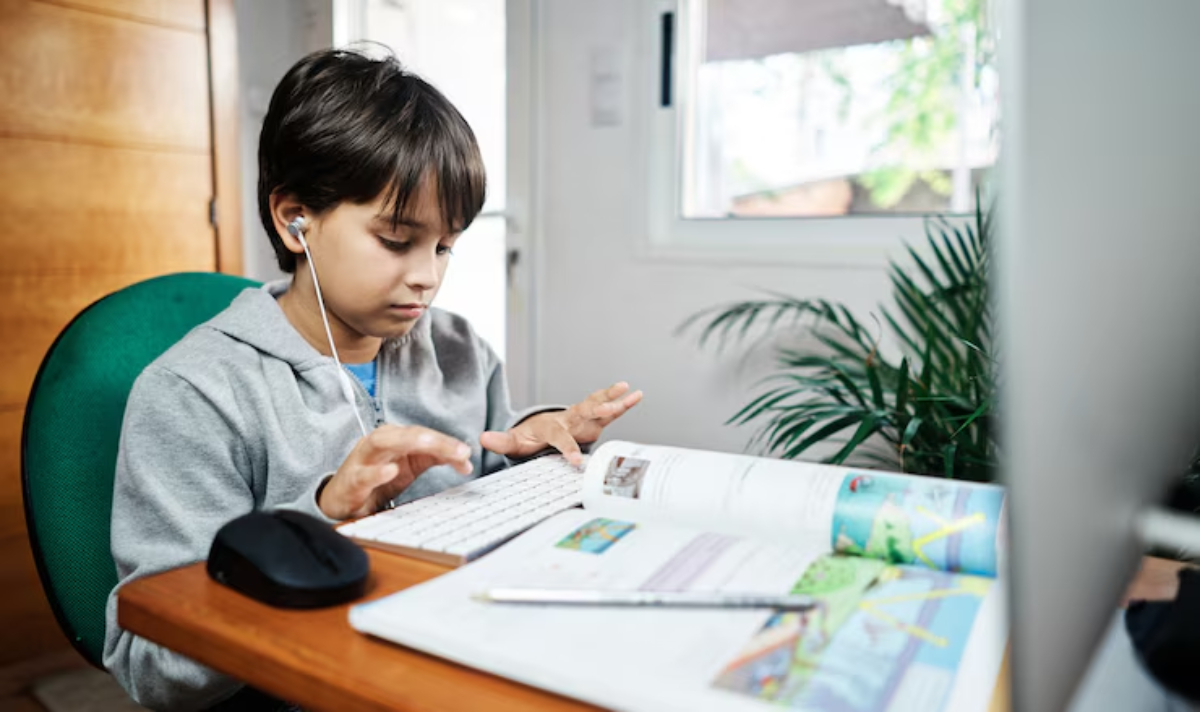 A child sits at a wooden desk engaged in online learning, with a keyboard, mouse, and open study materials beside them.
