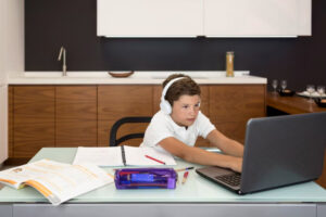 A child wearing headphones sits at a modern desk, focused on a laptop with notebooks and colored pens nearby.