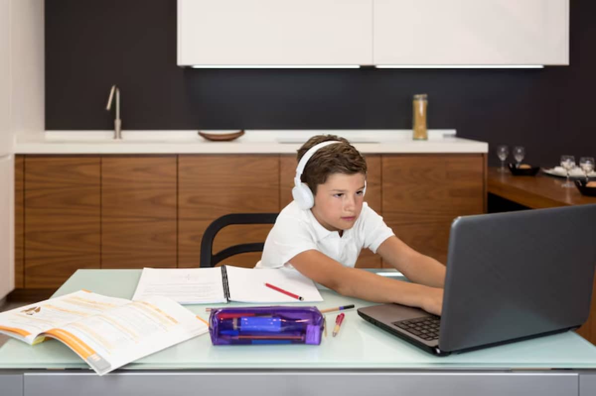 A child wearing headphones sits at a modern desk, focused on a laptop with notebooks and colored pens nearby.