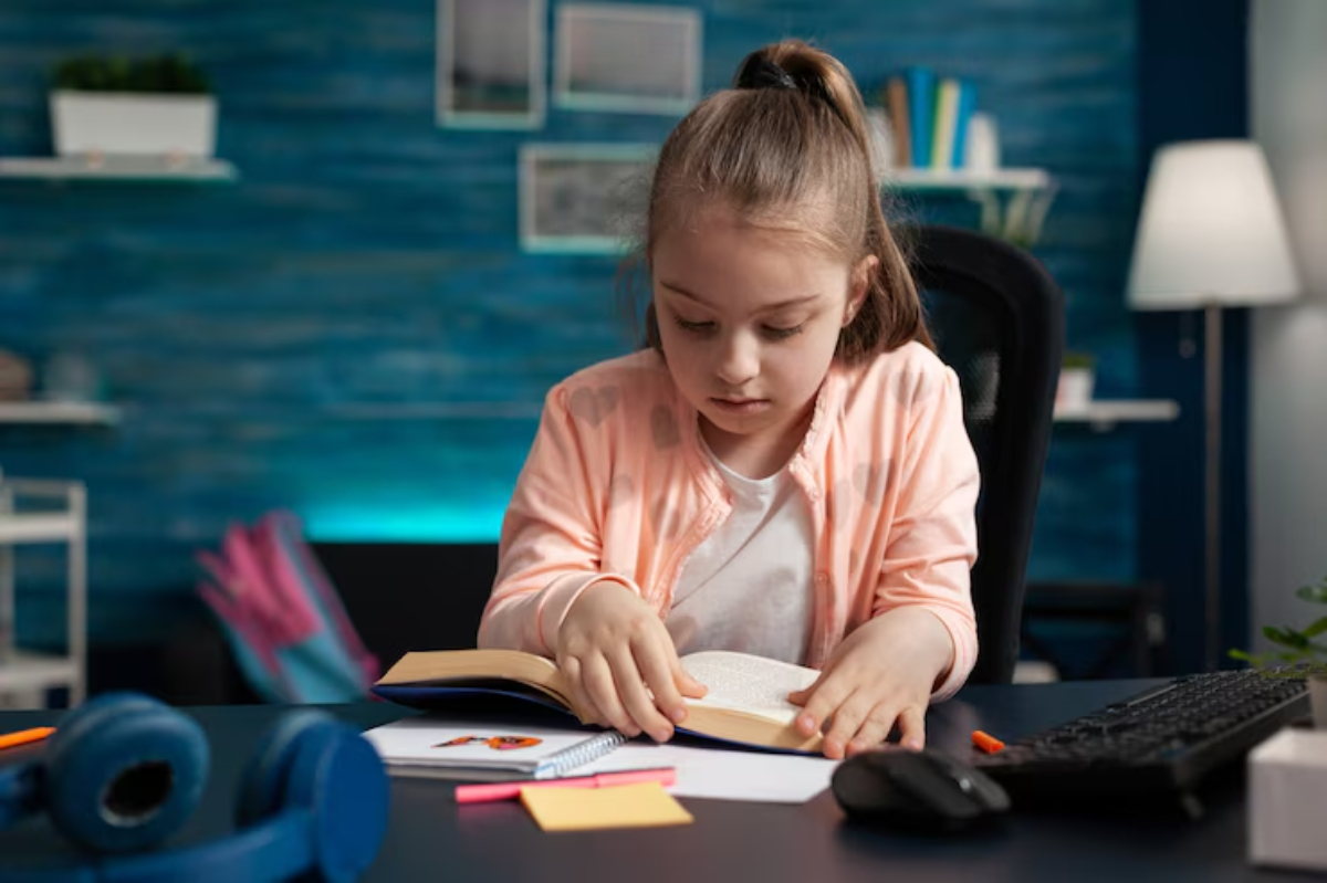A child studies at a desk, engaged with an open book, surrounded by notes, a notebook, headphones, and a computer keyboard.