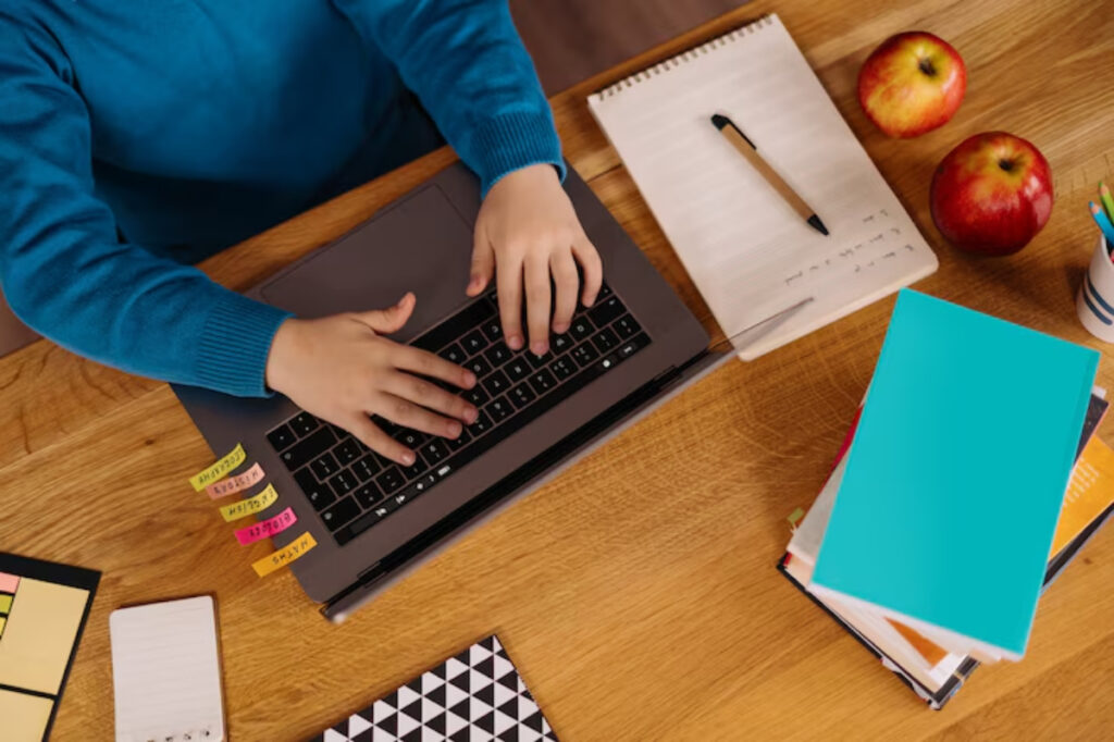 A person’s hands typing on a laptop at a wooden desk, surrounded by books, apples, sticky notes, and a notepad.