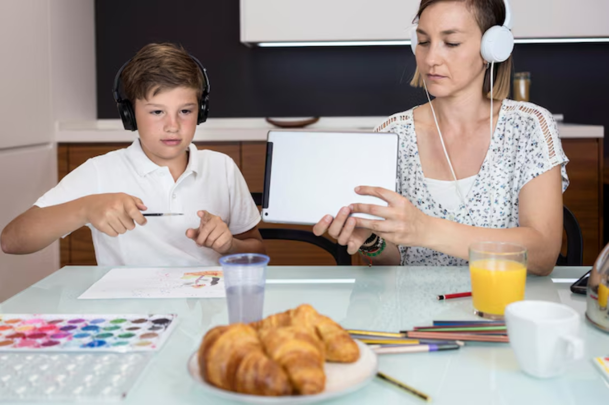 A child in headphones draws while an adult holds a tablet. Colorful crayons and croissants are on the table with drinks.