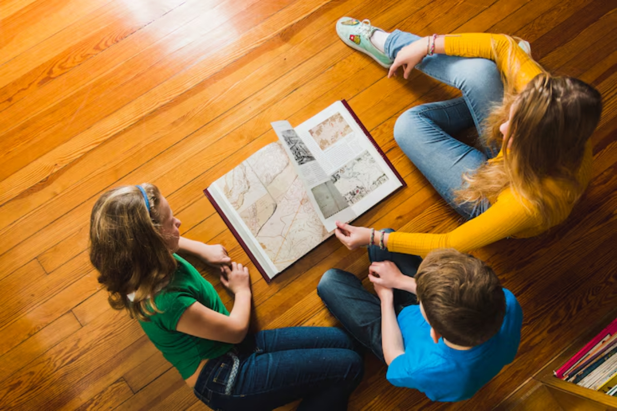 Three children sit on a wooden floor, examining a large book filled with maps and images, engaging in focused conversation.