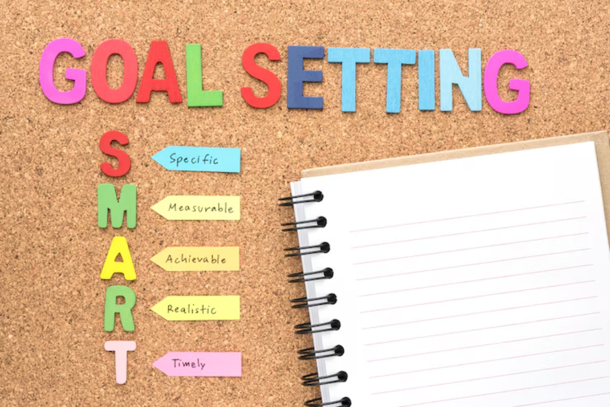 Colorful letters spell GOAL SETTING on a corkboard, with a list of SMART criteria and an empty notebook beside it.