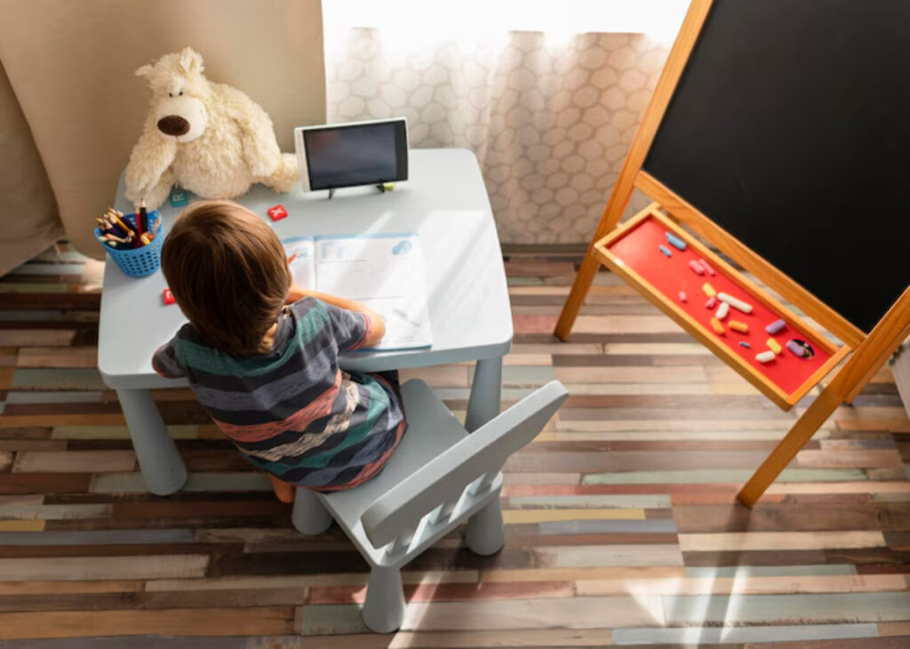 A young child with a striped shirt sits at a table, focused on homework, with a stuffed bear and a blackboard nearby.