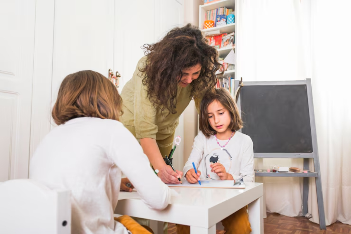 A woman assists two children in an art activity at a table, with bookshelves and a blackboard in a bright, cheerful room.