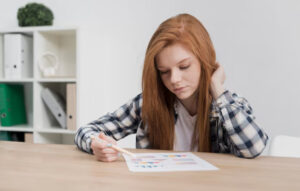 A person with long red hair sits at a wooden table, examining graphs and charts on a sheet of paper, looking contemplative.